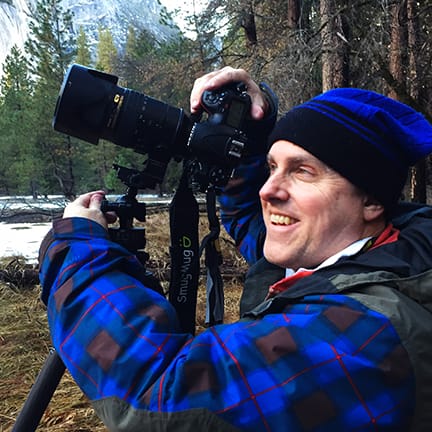 A landscape photographer using a Nikon DSLR on a Gitzo tripod while capturing winter scenery in Yosemite National Park, wearing a blue plaid jacket with snow-covered forest and granite cliffs in the background.