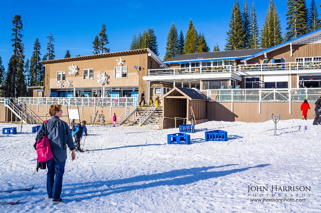 A wide view of the historic Badger Pass Day Lodge in Yosemite National Park with skiers and visitors enjoying a clear, sunny February day on the snow-covered slopes and outdoor sundeck.