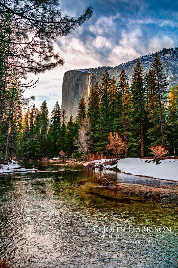 Winter sunset view of Horsetail Falls on El Capitan in Yosemite National Park, framed by pine trees and a reflective Merced River with snow along the riverbank.