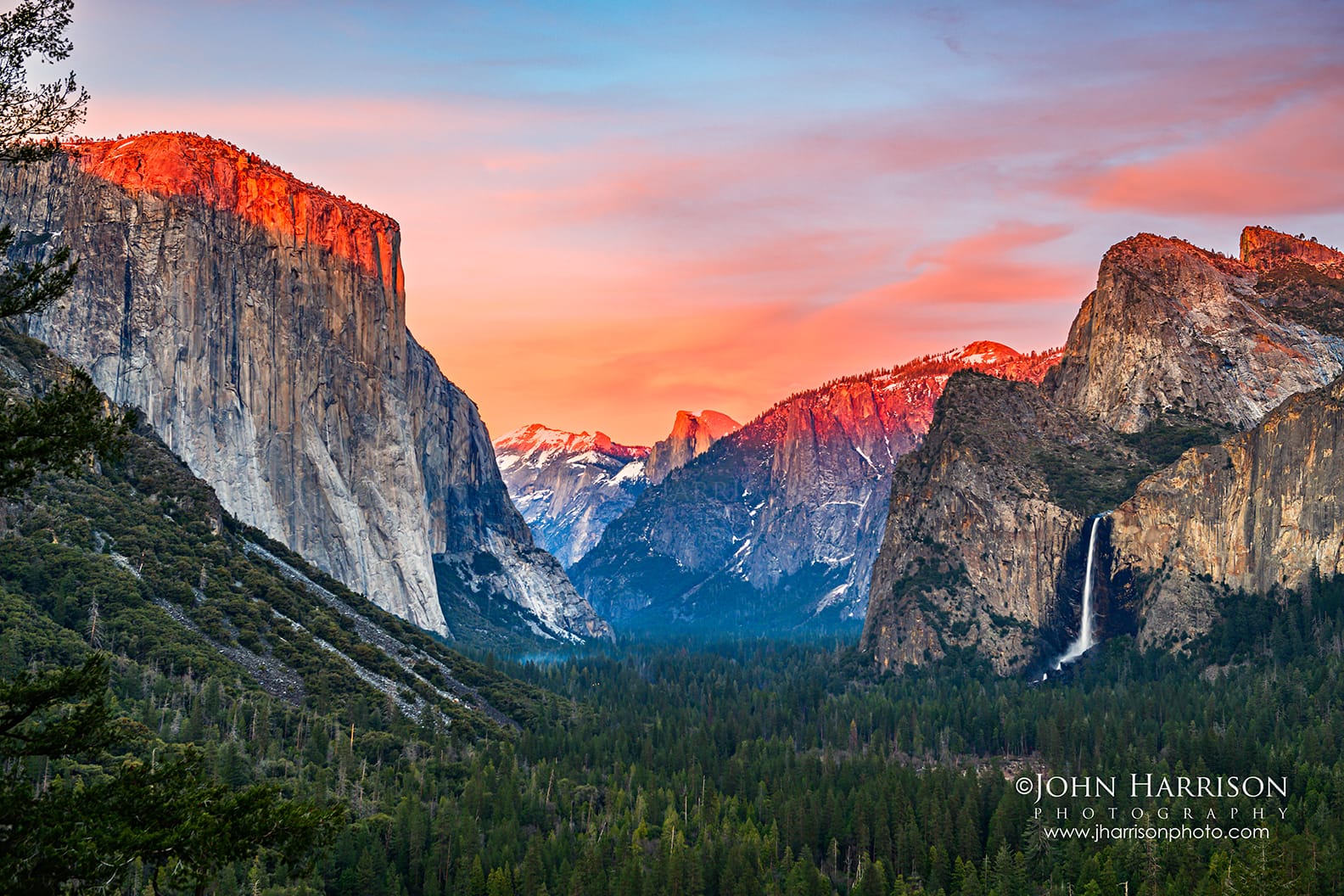Alpenglow illuminating El Capitan and Yosemite Valley at sunset from Tunnel View in Yosemite National Park, California, with Bridalveil Fall and dramatic spring clouds above the Sierra Nevada.
